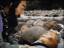 a man with a beard is standing next to a man laying on a rock