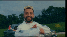 a man wearing sunglasses and a white shirt is holding a bag in front of a car