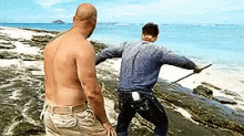 two shirtless men are standing on a rocky beach near the water