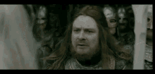 a man with long red hair and a beard is standing in front of a group of soldiers in armor .