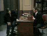 two men in suits are sitting at a desk in a library talking to each other .
