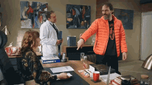 a man in an orange jacket is pointing at a woman sitting at a desk in front of a man in a lab coat .