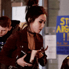 a woman in a brown jacket is standing in front of a sign that says stop arguing .