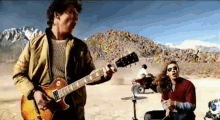 a man is playing a guitar in the desert while another man sings