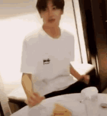 a man in a white t-shirt is sitting at a table with a plate of food .