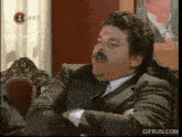 a man in a suit and tie with a mustache is sitting in a chair with a gifrun.com logo in the corner