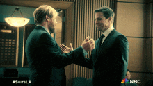 two men in suits shake hands in front of a sign that says nbc on it
