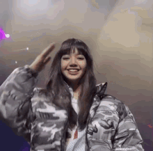 a woman in a camouflage jacket is smiling and waving her hand .