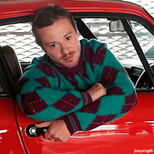 a man in a green and purple plaid sweater leans out of a red car