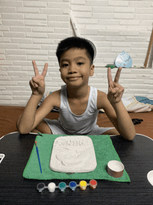 a young boy gives a peace sign while sitting at a table with paints