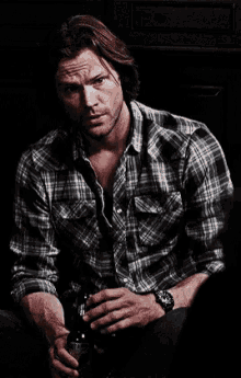 a man in a plaid shirt is sitting in a dark room with a bottle of beer in his hand .