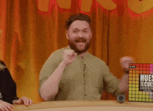 a man with a beard is sitting at a table with his fist in the air in front of a curtain .