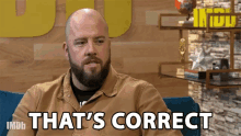 a bald man with a beard is sitting on a blue couch and saying that 's correct .