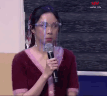 a woman wearing a face shield and glasses is holding a microphone in front of her face .