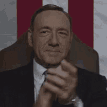 a man in a suit and tie is clapping his hands while sitting in front of a flag .