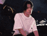 a man wearing a white shirt with the letter t on the back sits in a chair