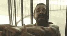 a man without a shirt is behind bars in a jail cell .