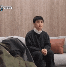 a man in a black sweater is sitting on a couch in front of a wooden wall .
