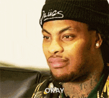 a man wearing a beanie and earrings is sitting on a couch and saying okay .
