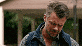 a man with a beard wearing a plaid shirt and a vest is standing in front of a house .