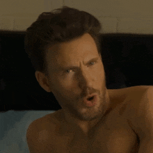 a shirtless man with a beard is laying on a bed and making a face .