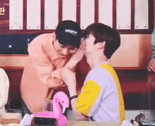 a man is kissing another man on the cheek while sitting at a table with a pink flamingo .