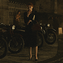 a woman in a black dress stands in front of a motorcycle and a fence