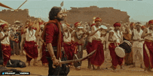 a man holding a sword stands in front of a group of men playing drums and drums
