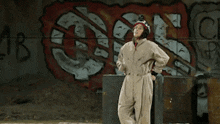 a woman in a jumpsuit is standing in front of a wall with graffiti on it including the letters ab and c