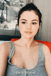 a woman in a grey tank top is sitting in front of a window with the words okie dokie below her