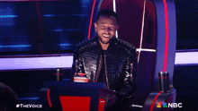 a man in a leather jacket is sitting in a chair with a nbc logo behind him