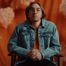 a man in a denim jacket and glasses sits in a chair
