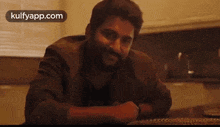 a man with a beard is sitting at a table in a kitchen talking to someone .
