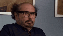 a bald man with a beard and glasses is sitting in front of a picture .