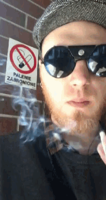a man wearing sunglasses and a hat smoking a cigarette in front of a no smoking sign