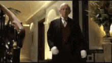 a man in a tuxedo and white gloves is standing in a hallway next to a staircase .