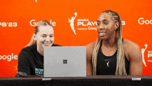 two women are looking at a laptop in front of a wall that says wnba play
