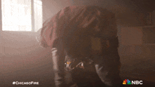 a man in a red jacket is kneeling down in a room with a nbc logo in the corner