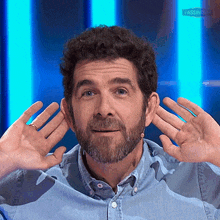 a man with a beard is covering his ears with his hands while wearing a blue shirt