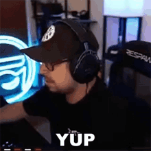a man wearing headphones and a hat is sitting in front of a computer and says yup .