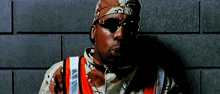 a man wearing sunglasses and a camouflage jacket stands in front of a wall
