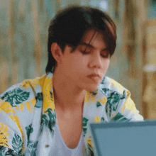 a young man wearing a floral shirt is sitting in front of a laptop .