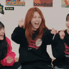 a woman with red hair is laughing while sitting next to two other women in front of a wall with basketball posters on it .