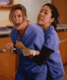 two women in blue scrubs are hugging each other in a hospital room .