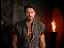 a man with a beard wearing a leather vest is standing in front of a fire .