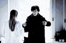 a man in a black coat is standing next to a woman in a white shirt in front of a window .