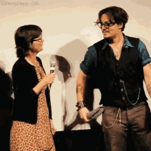 a woman is holding a microphone and talking to a man who is wearing glasses and a black vest