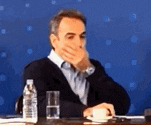a man is covering his mouth with his hand while sitting at a table with a cup of coffee and a bottle of water .