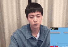 a young man in a denim jacket is sitting in front of a computer screen and making a funny face .