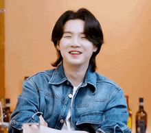 a young man wearing a denim jacket is smiling while sitting at a table with bottles in the background .